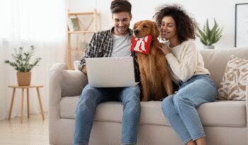 Smiling couple with their golden retriever holding a red toy while using a laptop together at home, representing shared pet custody after divorce.