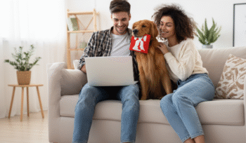 Smiling couple with their golden retriever holding a red toy while using a laptop together at home, representing shared pet custody after divorce.