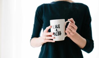 Women in an oversized black top with a white background holding a coffee mug in both hands that says, Like A Boss