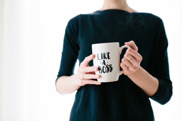 Women in an oversized black top with a white background holding a coffee mug in both hands that says, Like A Boss