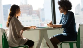 A couple sitting at a pedestal table having a casual conversation next to a window willed with sunlight