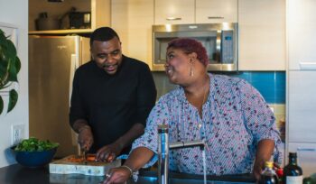 A happy, heavy set black couple helping each other in a kitchen sink cutting up tomatos.