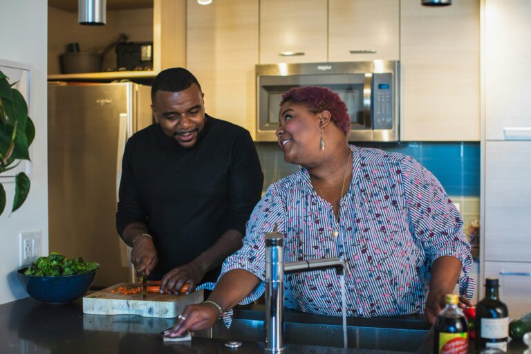 A happy, heavy set black couple helping each other in a kitchen sink cutting up tomatos.