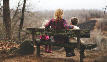 Mother sitting between 2 young children with arms around them on a rustic wooden bench overlooking an expanse of a forest