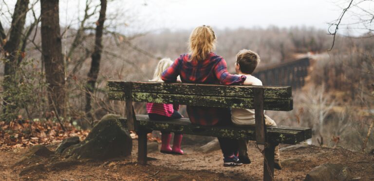 Mother sitting between 2 young children with arms around them on a rustic wooden bench overlooking an expanse of a forest