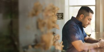 a man in a blue shirt stands at a wooden bar by a window in a cafe while he looks at his phone in contemplation