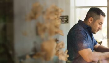 a man in a blue shirt stands at a wooden bar by a window in a cafe while he looks at his phone in contemplation